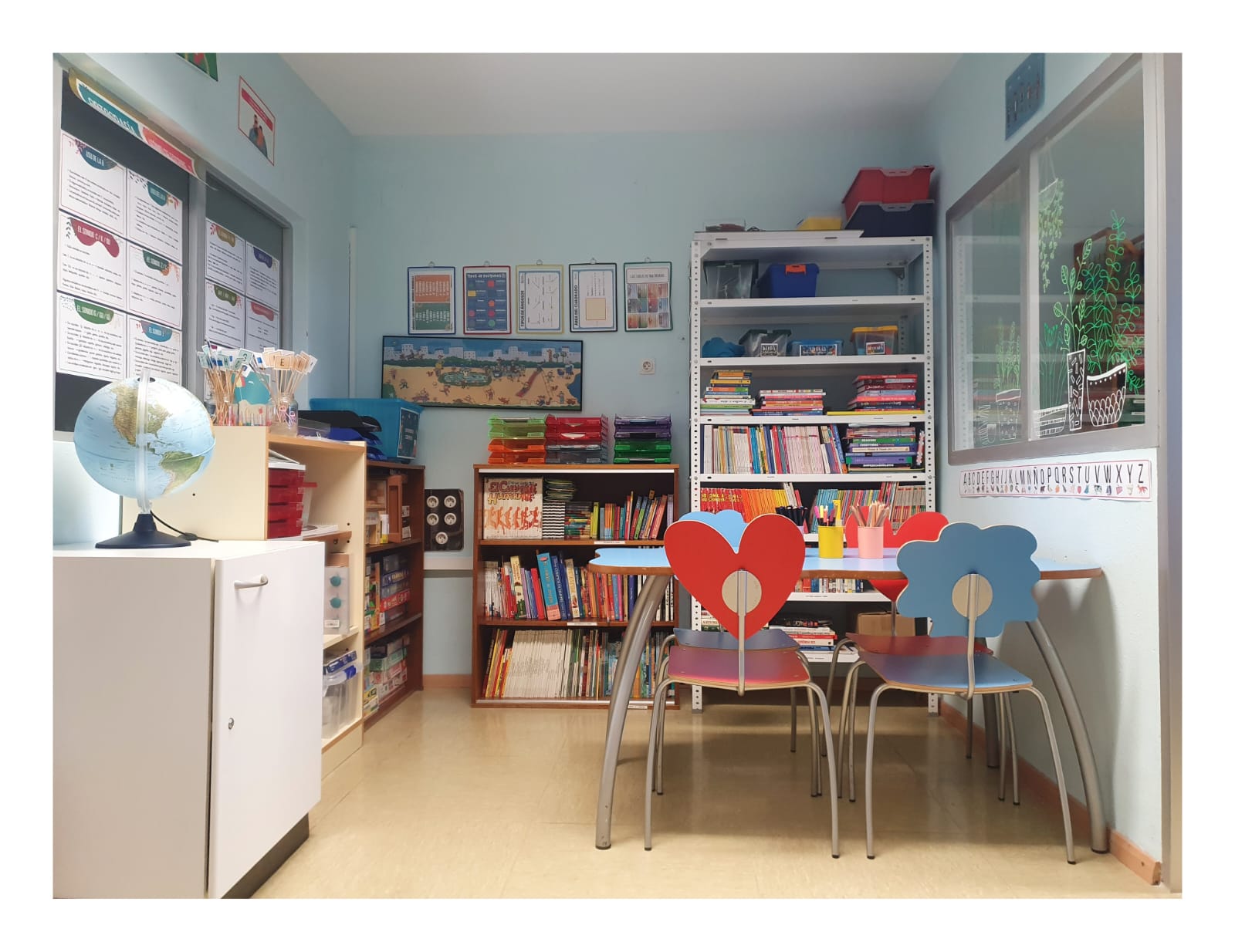 Aula interior de vistosos colores, con un escritorio, 4 sillas y gran cantidad de material escolar diverso tanto lectivo como para realizar actividades