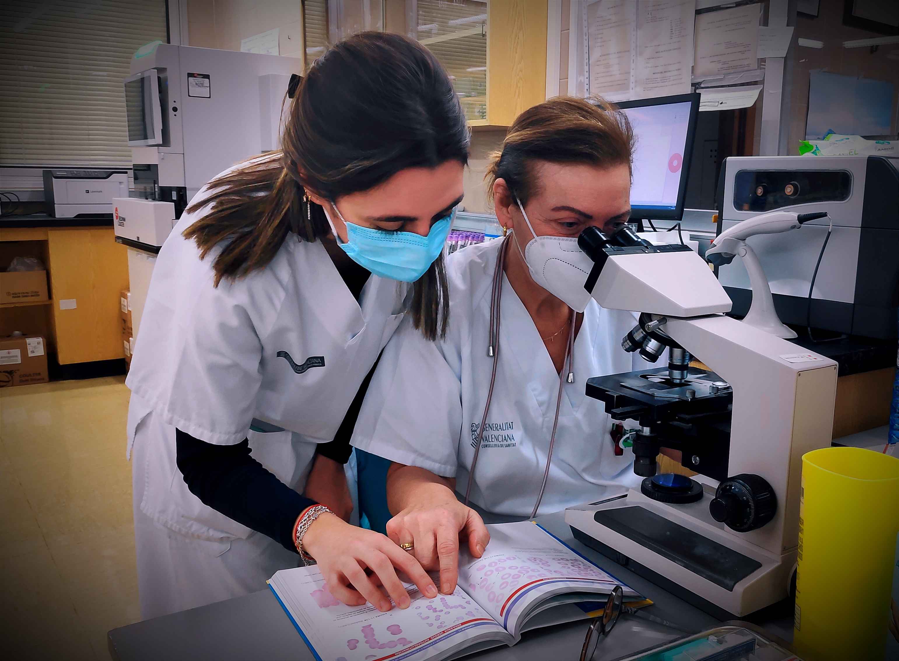 Sanitaria analizando muestras en un microscopio mientras compara junto a una compañera unas imágenes de un libro 